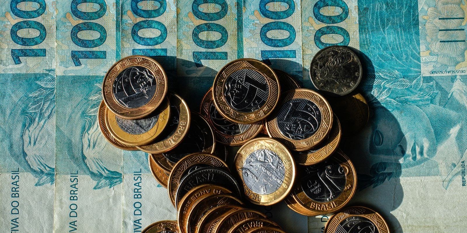 Notas de R$ 100 e moedas de R$ 1 espalhadas sobre a mesa