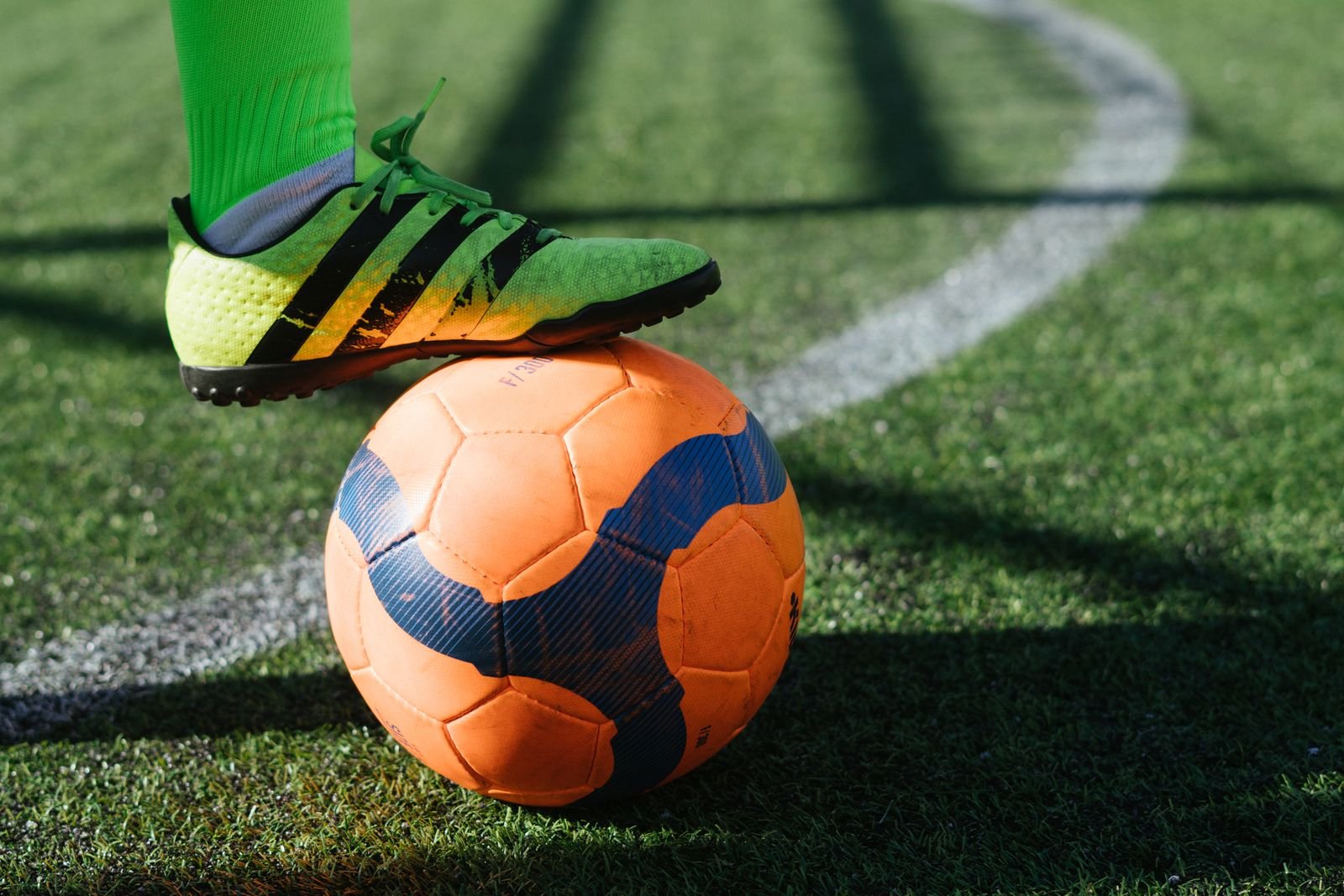 Pé de jogador com chuteira verde sobre uma bola laranja em campo de grama sintética