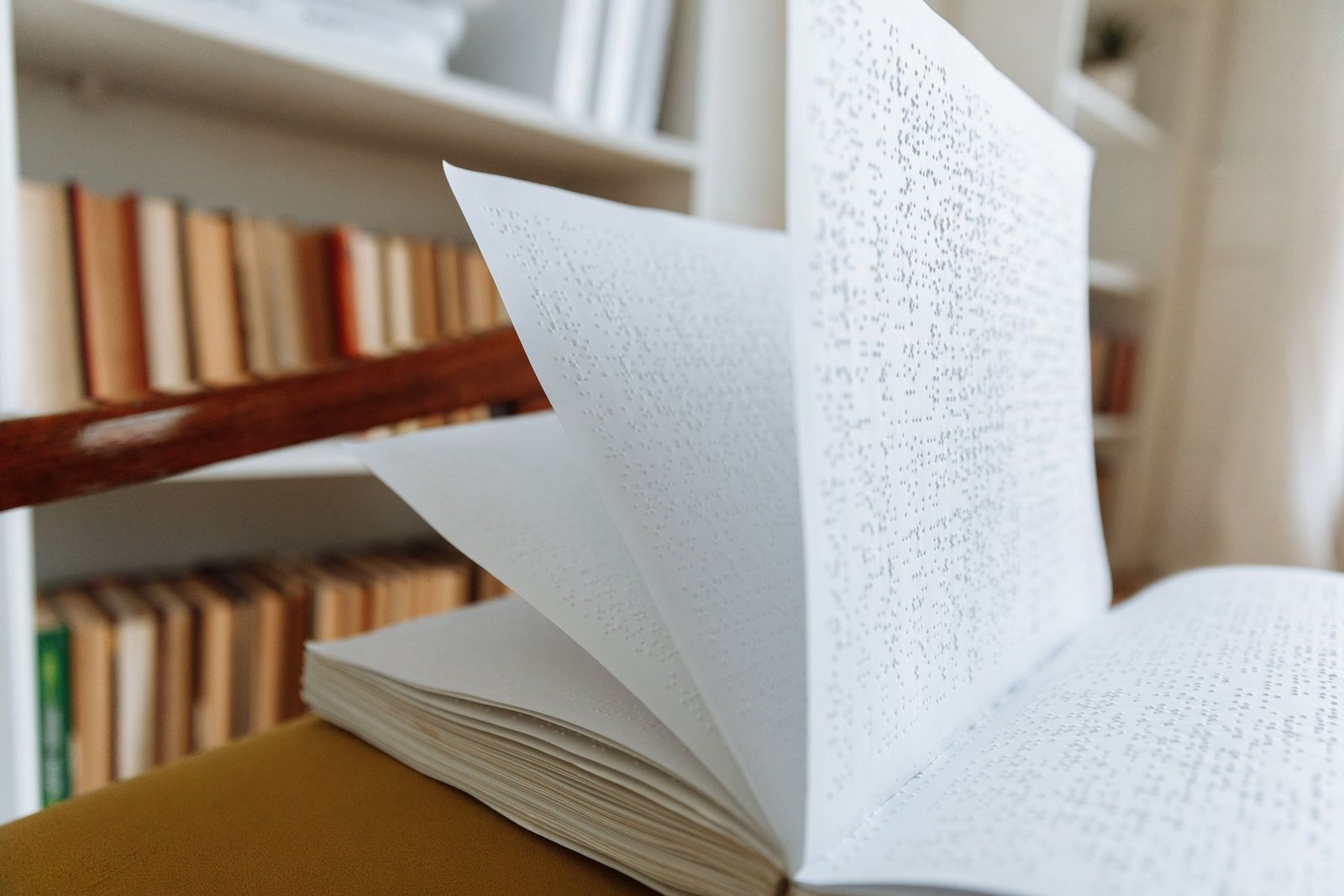 Livro em braile aberto, com páginas em movimento, diante de uma estante com livros (foto ilustrativa)