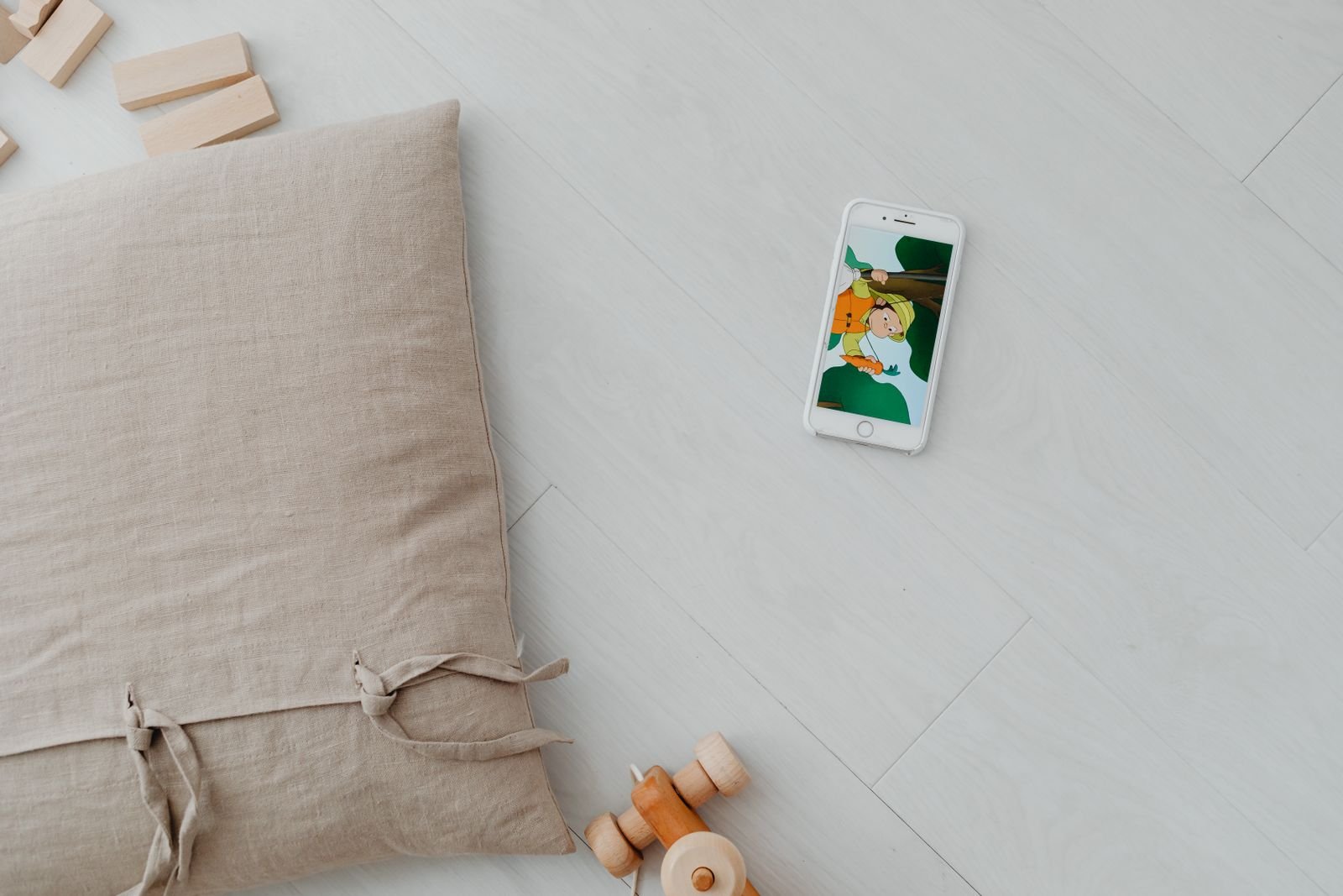 Celular com desenho animado sobre o piso, ao lado de brinquedos de madeira e uma almofada; foto ilustrativa
