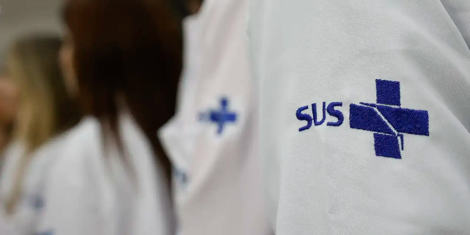 Detalhe do jaleco de profissionais de saúde com o símbolo do SUS bordado na manga