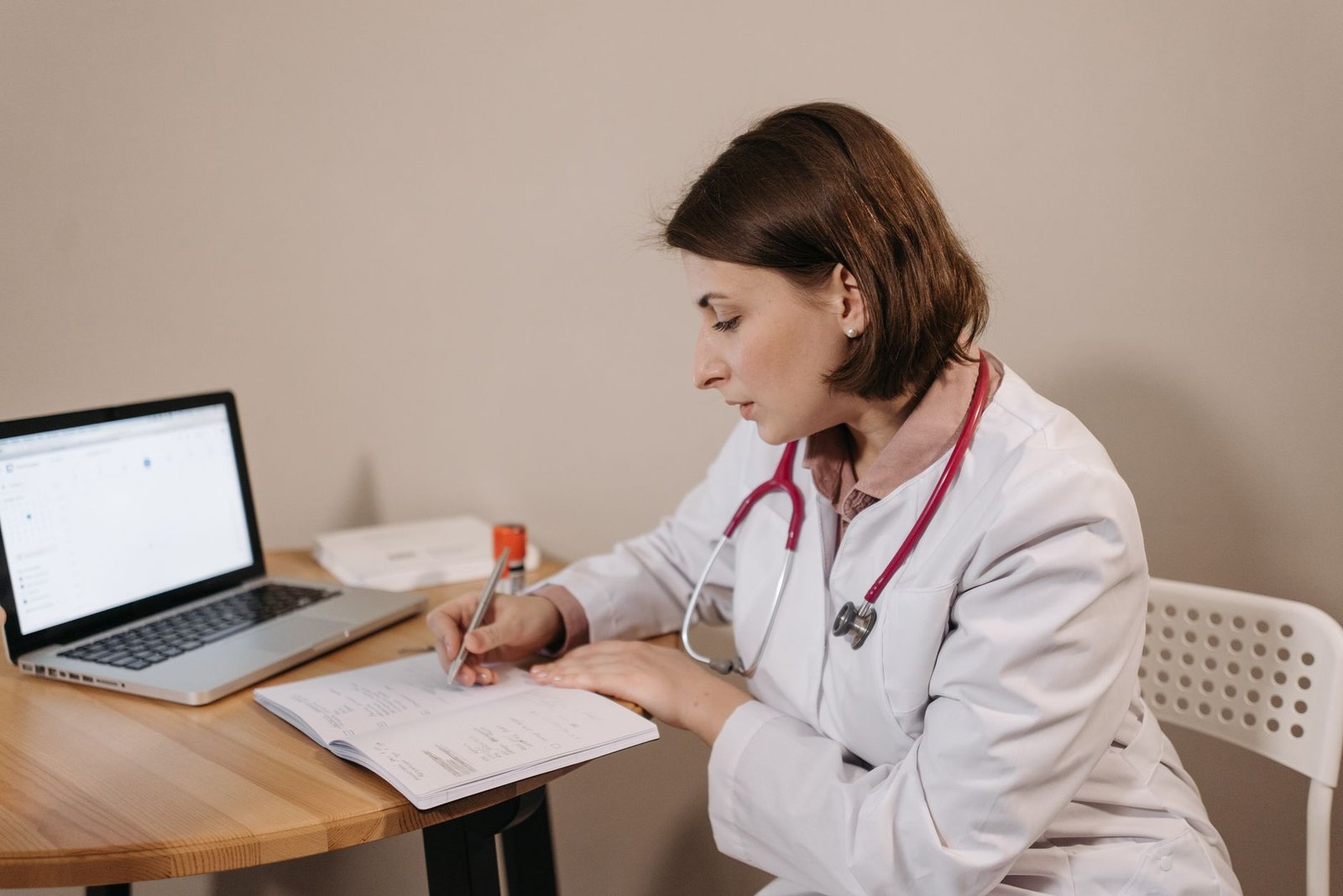 Médica de jaleco e estetoscópio escreve em um caderno diante de um notebook, em consultório