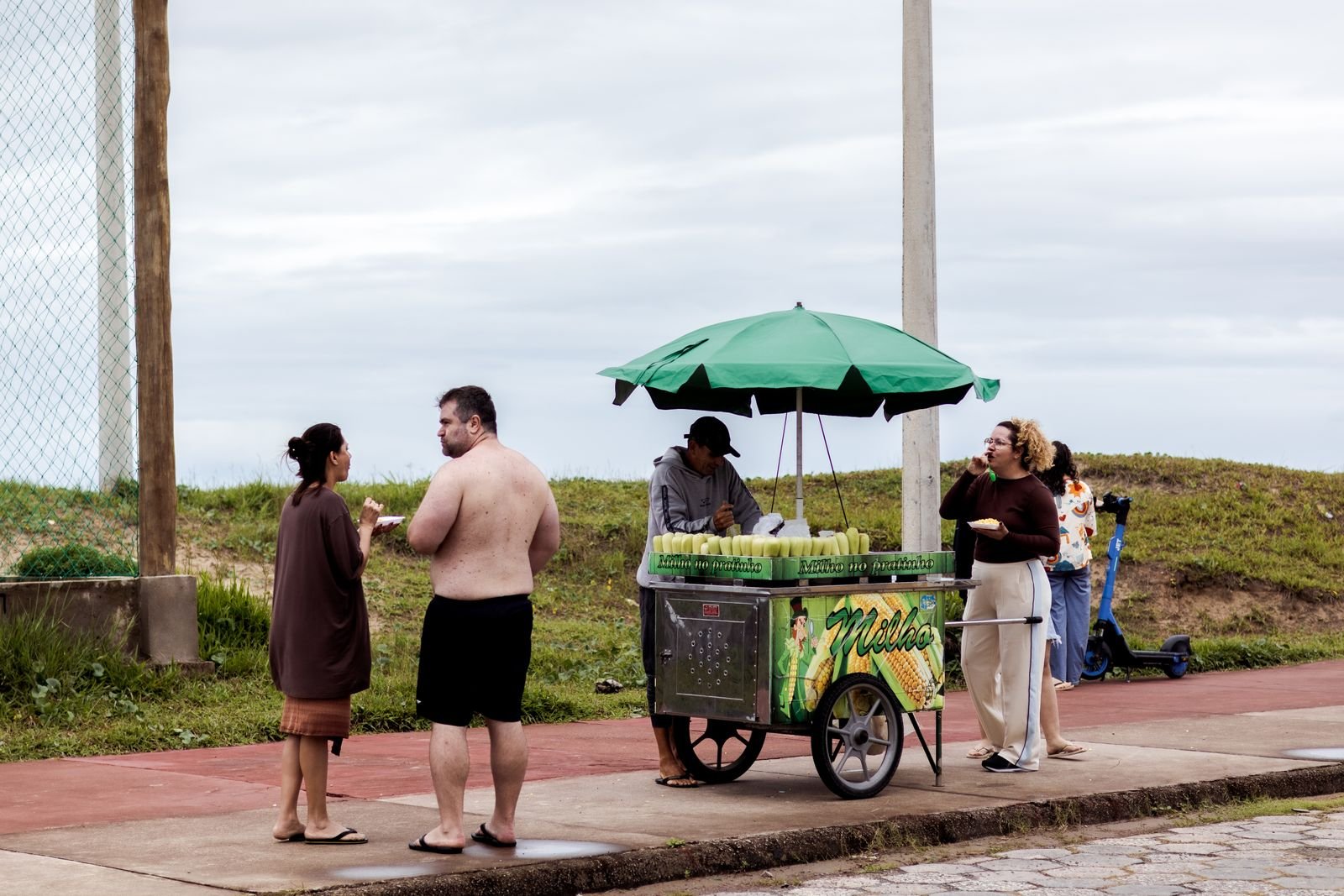 Vendedor de milho ambulante atende clientes em calçada à beira-mar, sob guarda-sol verde