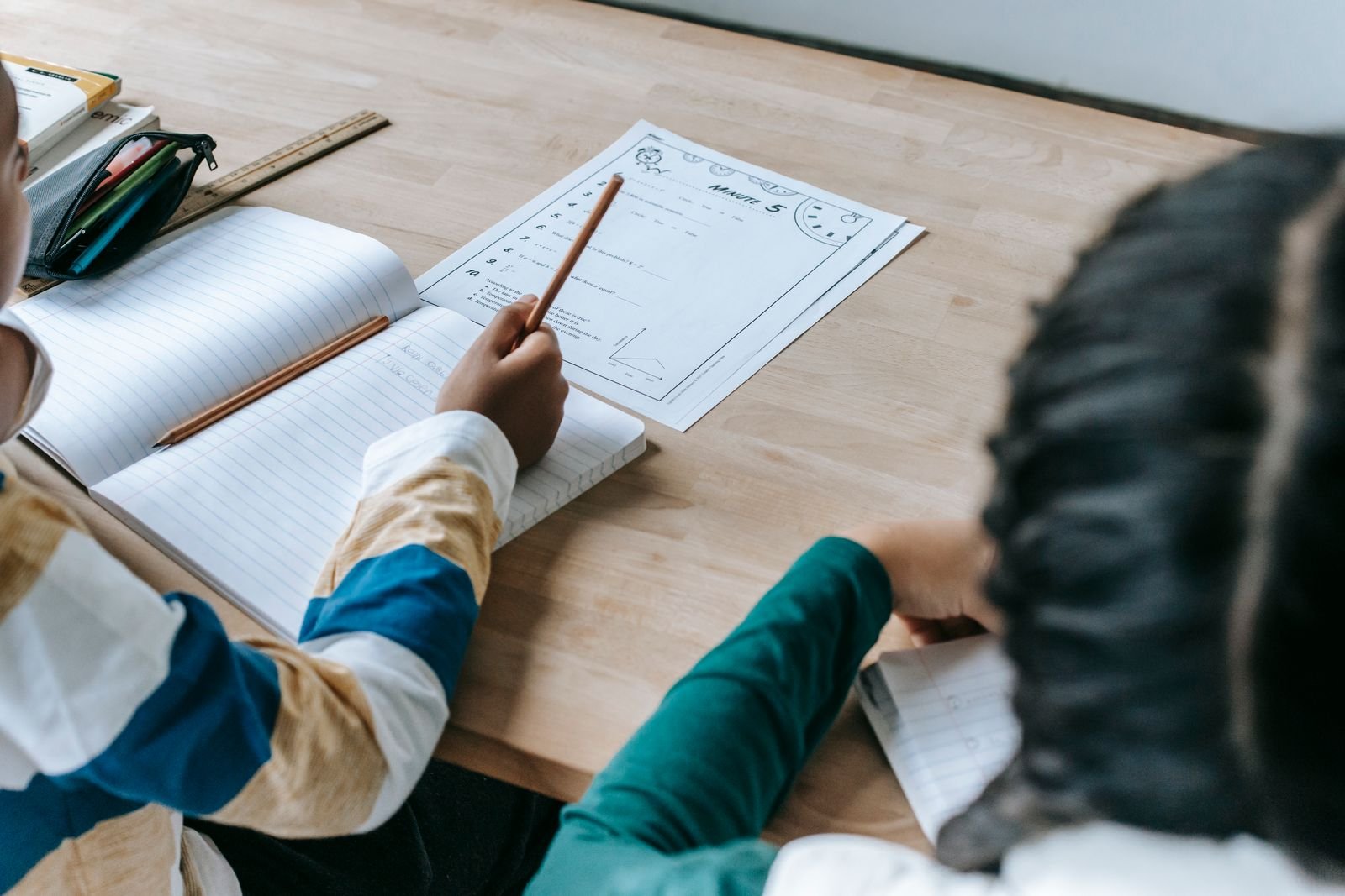 Estudantes escrevem em cadernos sobre uma mesa de madeira, imagem ilustrativa