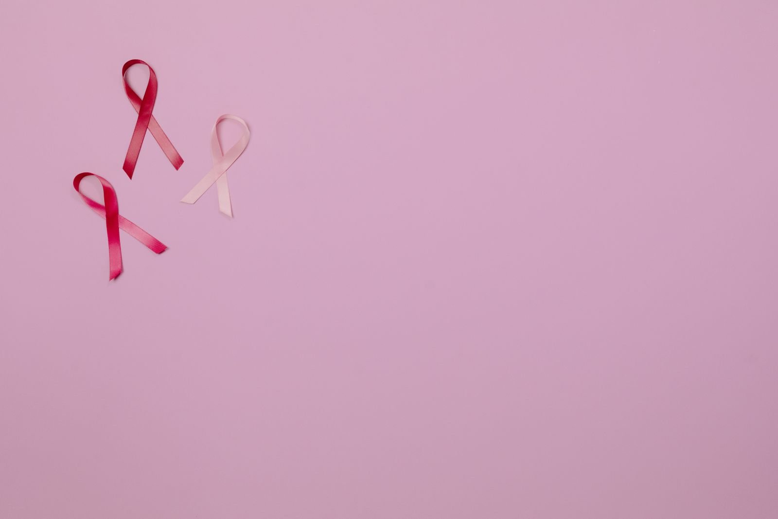 Três laços de fita nas cores rosa e vermelho sobre fundo rosa, símbolo da campanha Outubro Rosa