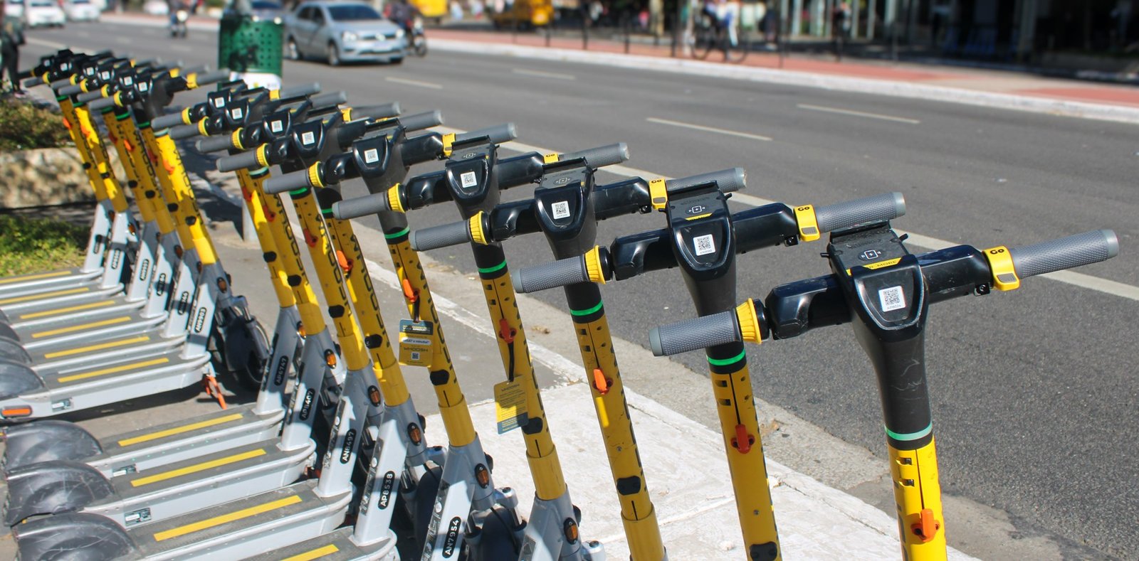 Fileira de patinetes elétricos amarelos de aluguel estacionados na calçada de uma avenida (foto de arquivo, 2025)