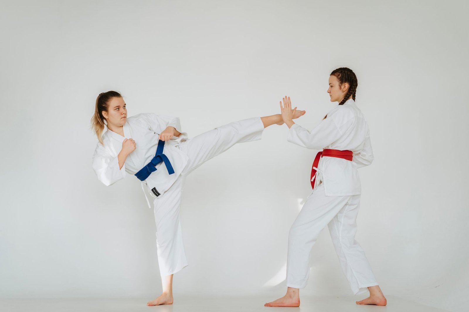 Duas mulheres de quimono branco treinam artes marciais em estúdio; uma aplica um chute alto e a outra defende com a mão