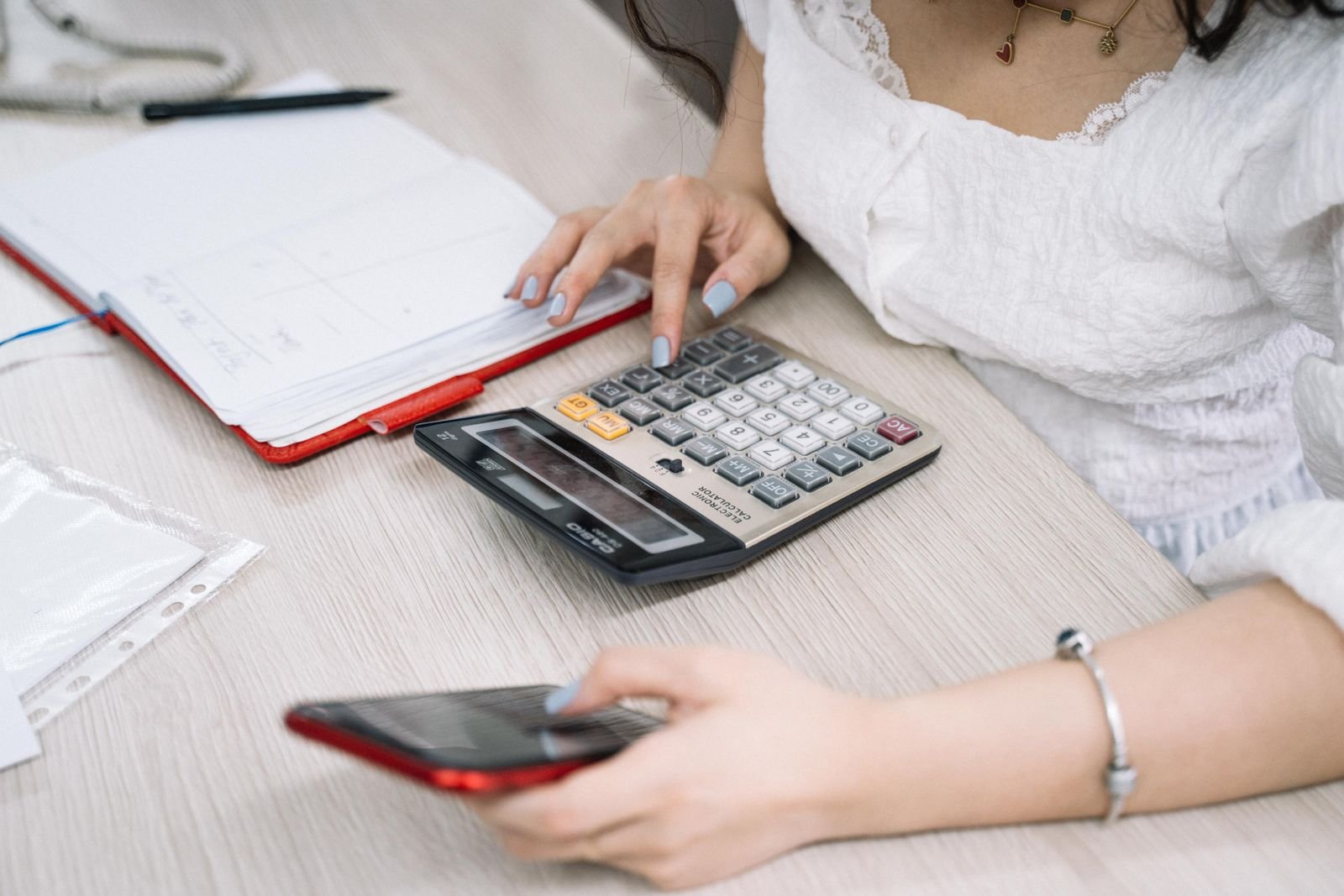 Mulher digita em calculadora sobre mesa de escritório, com caderno e celular ao lado (foto ilustrativa)