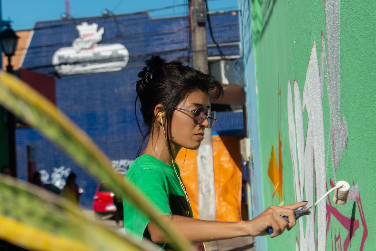 Artista pinta com rolo um mural em parede verde numa rua de bairro