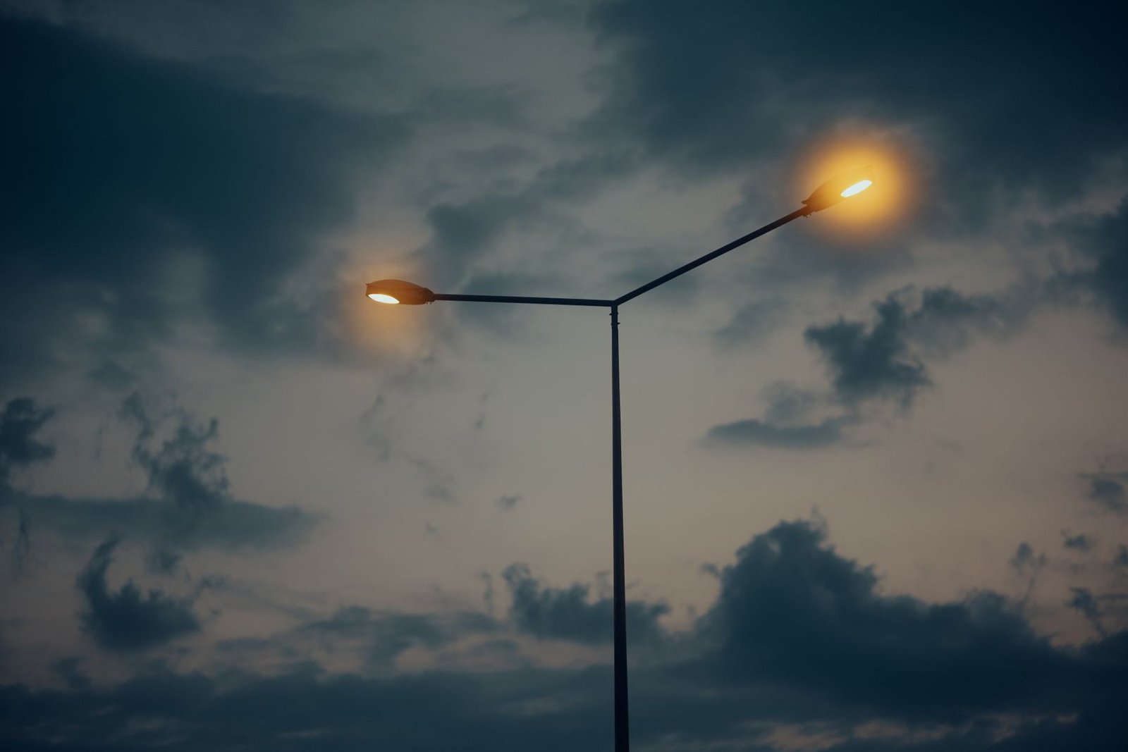 Poste de iluminação pública com duas luminárias acesas ao entardecer, contra céu com nuvens