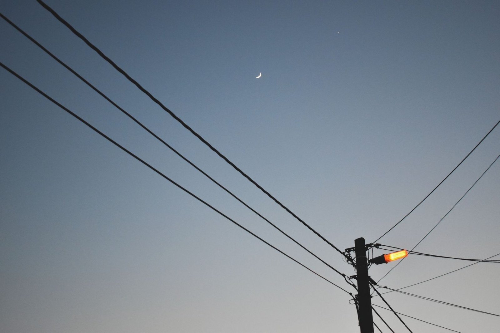 Poste de iluminação pública com luminária acesa ao entardecer, entre fios de rede, com a lua crescente ao fundo