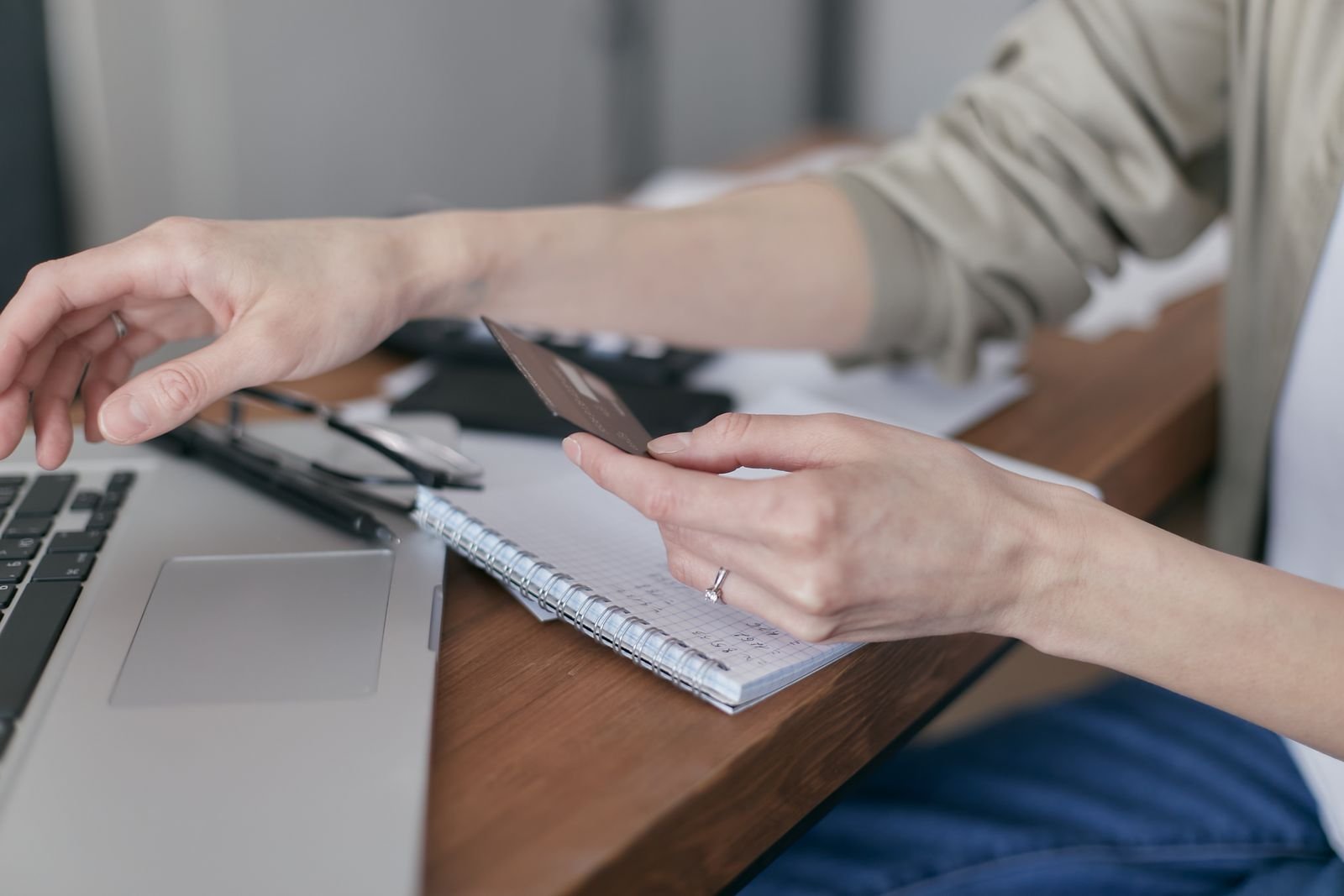 Mulher segura um cartão enquanto usa notebook sobre mesa com caderno de anotações