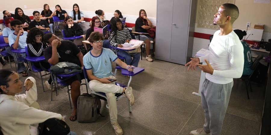 Professor de pé à frente de uma sala de aula com alunos sentados em carteiras