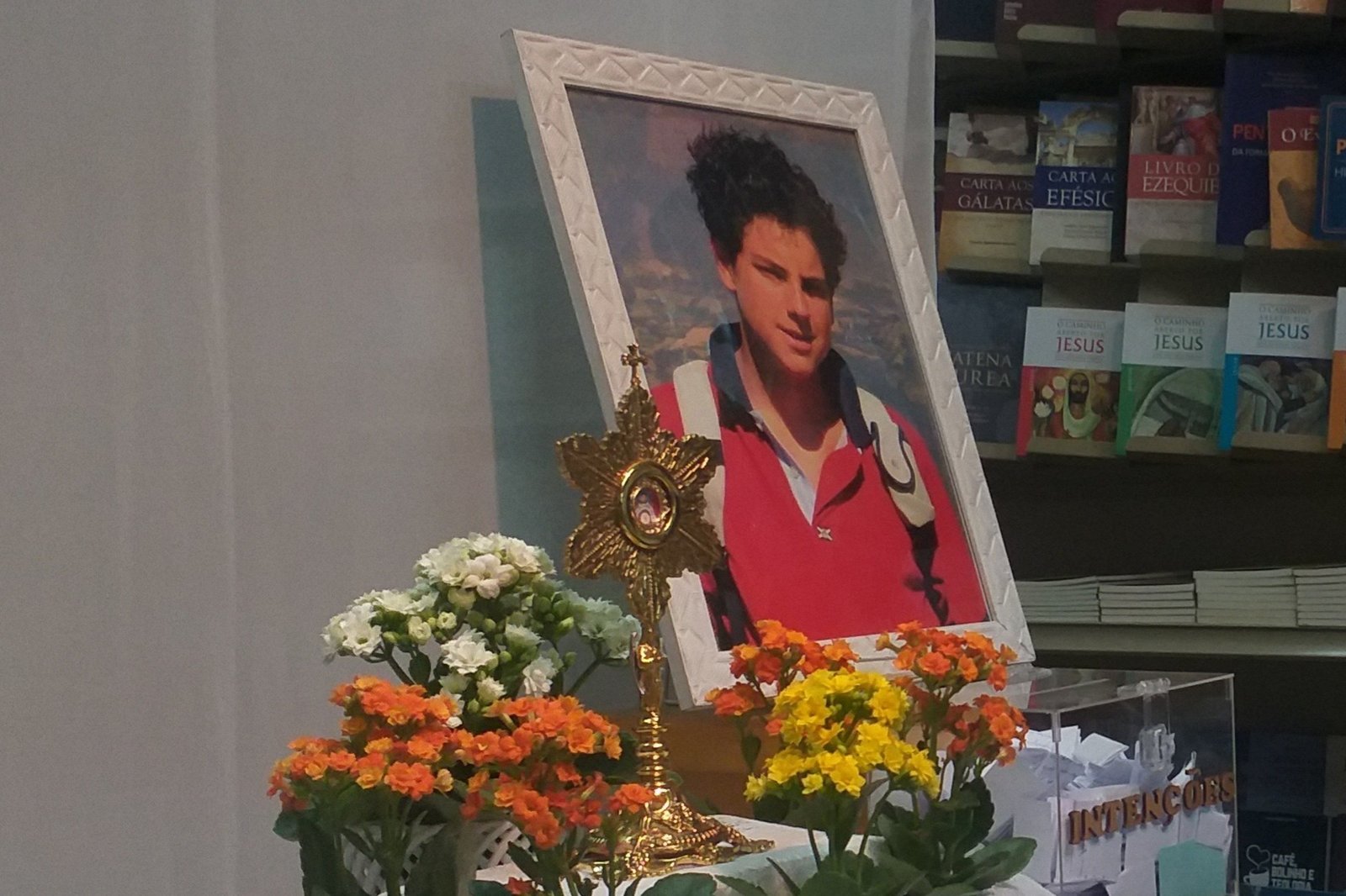 Relicário dourado diante de retrato de Carlo Acutis, com flores e a placa Não tocar na relíquia