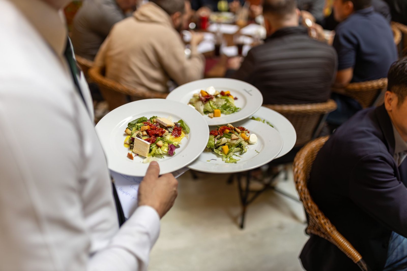 Foto ilustrativa de garçom carregando pratos de salada em restaurante com clientes sentados às mesas