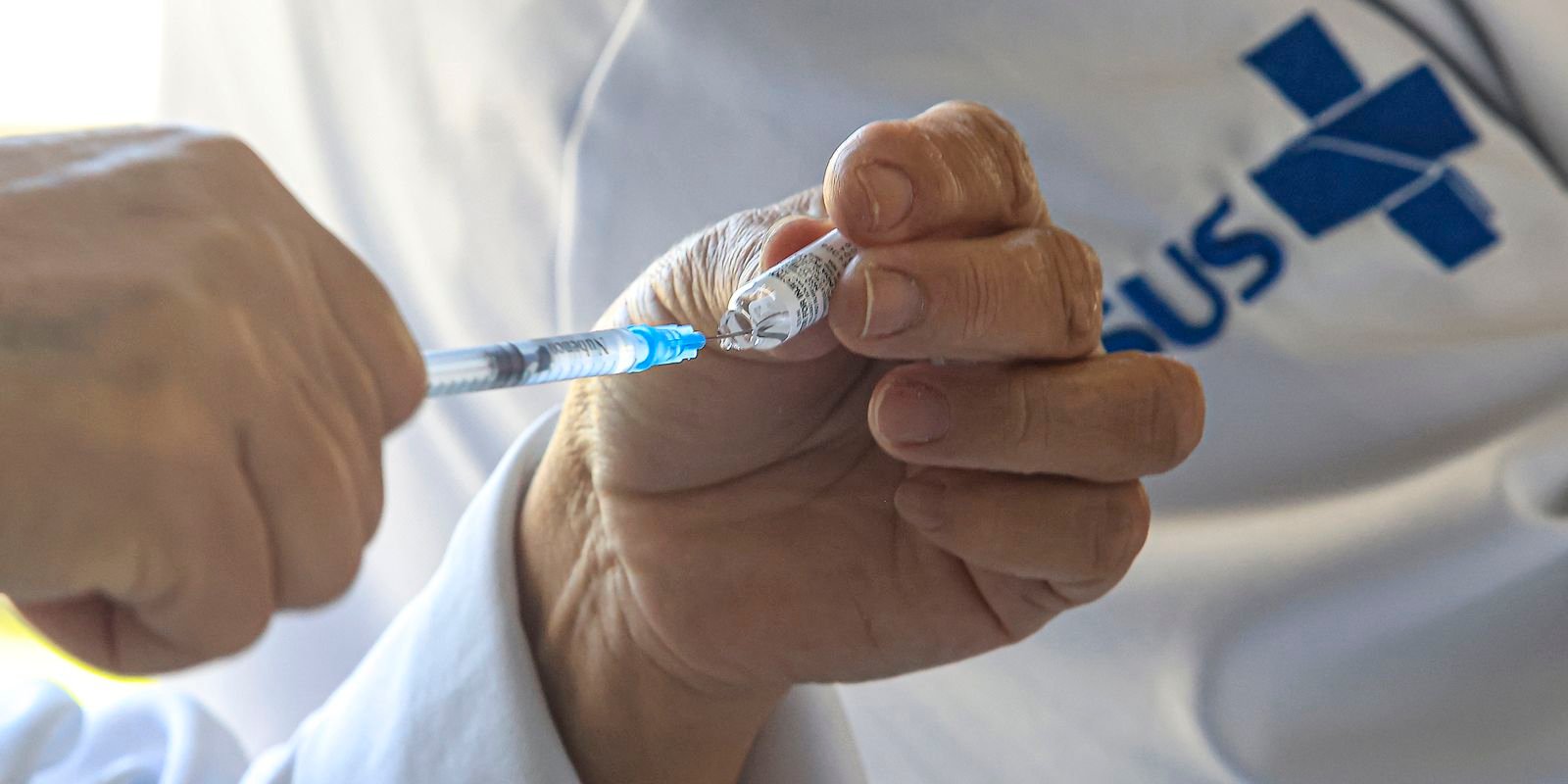 Profissional de saúde prepara a seringa com a vacina, com a marca do SUS ao fundo