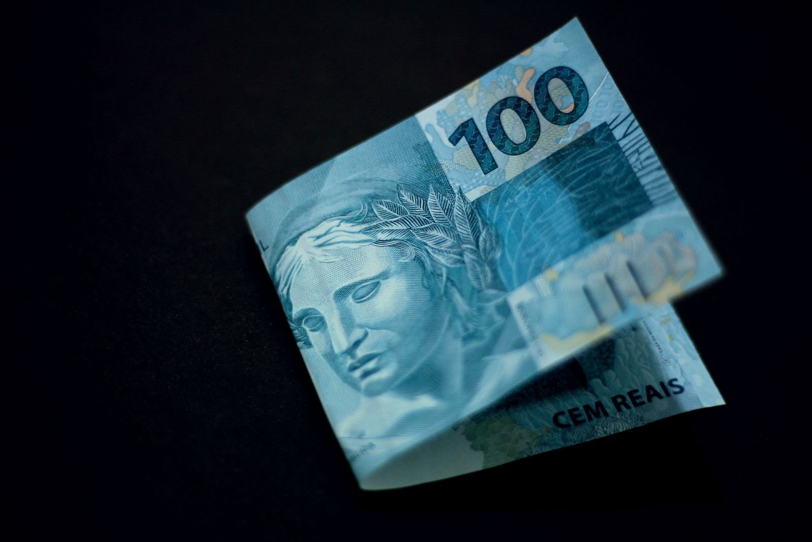 Cédula de 100 reais sobre fundo escuro, imagem ilustrativa