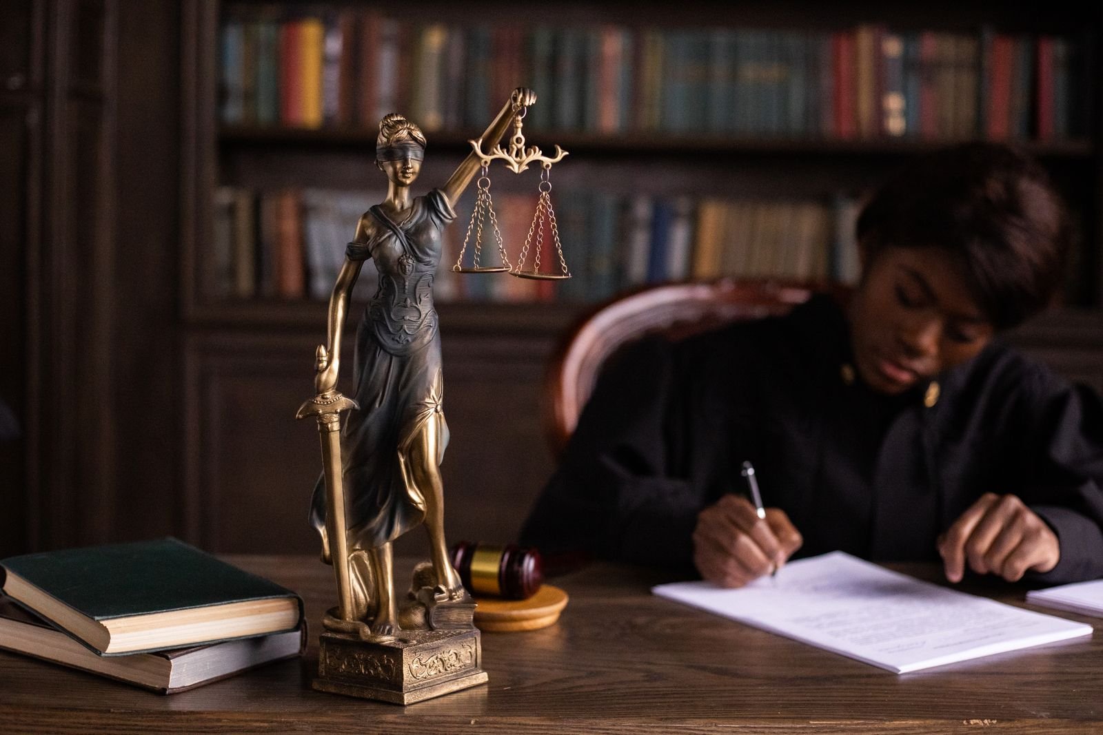 Estátua da Justiça sobre a mesa, com livros e martelo, e uma juíza assinando documentos ao fundo
