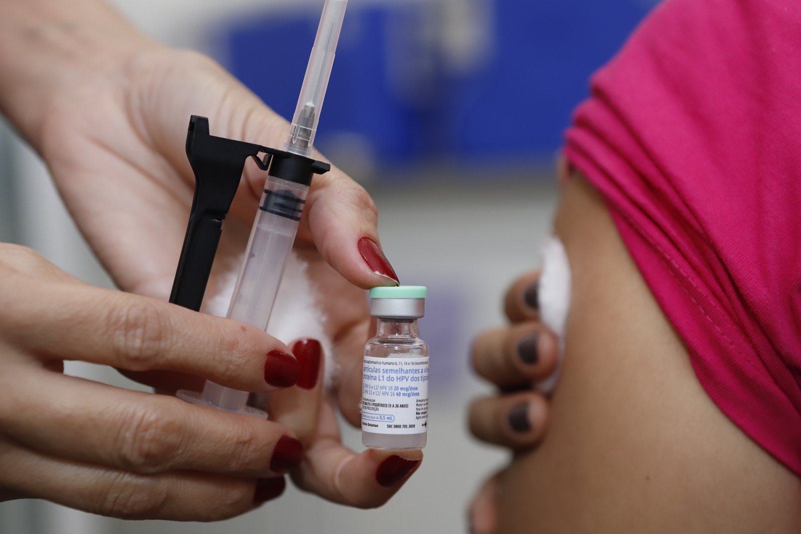 Mãos de uma profissional de saúde seguram um frasco da vacina contra HPV e uma seringa durante aplicação no braço de uma paciente