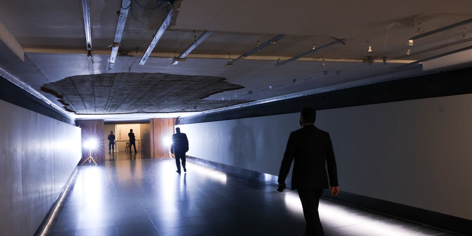 Corredor subterrâneo da Câmara dos Deputados com um buraco no teto onde o reboco caiu; duas pessoas caminham pelo piso iluminado
