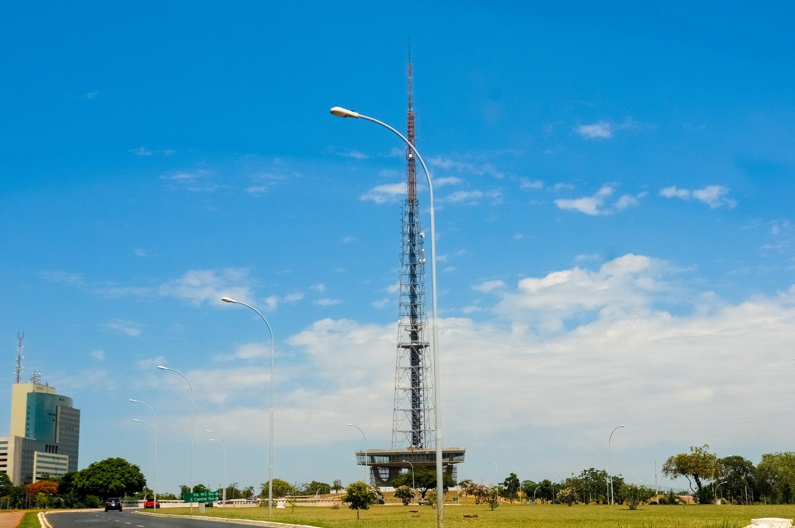 Torre de TV de Brasília ao fundo, com céu azul, gramado e via do Eixo Monumental em primeiro plano