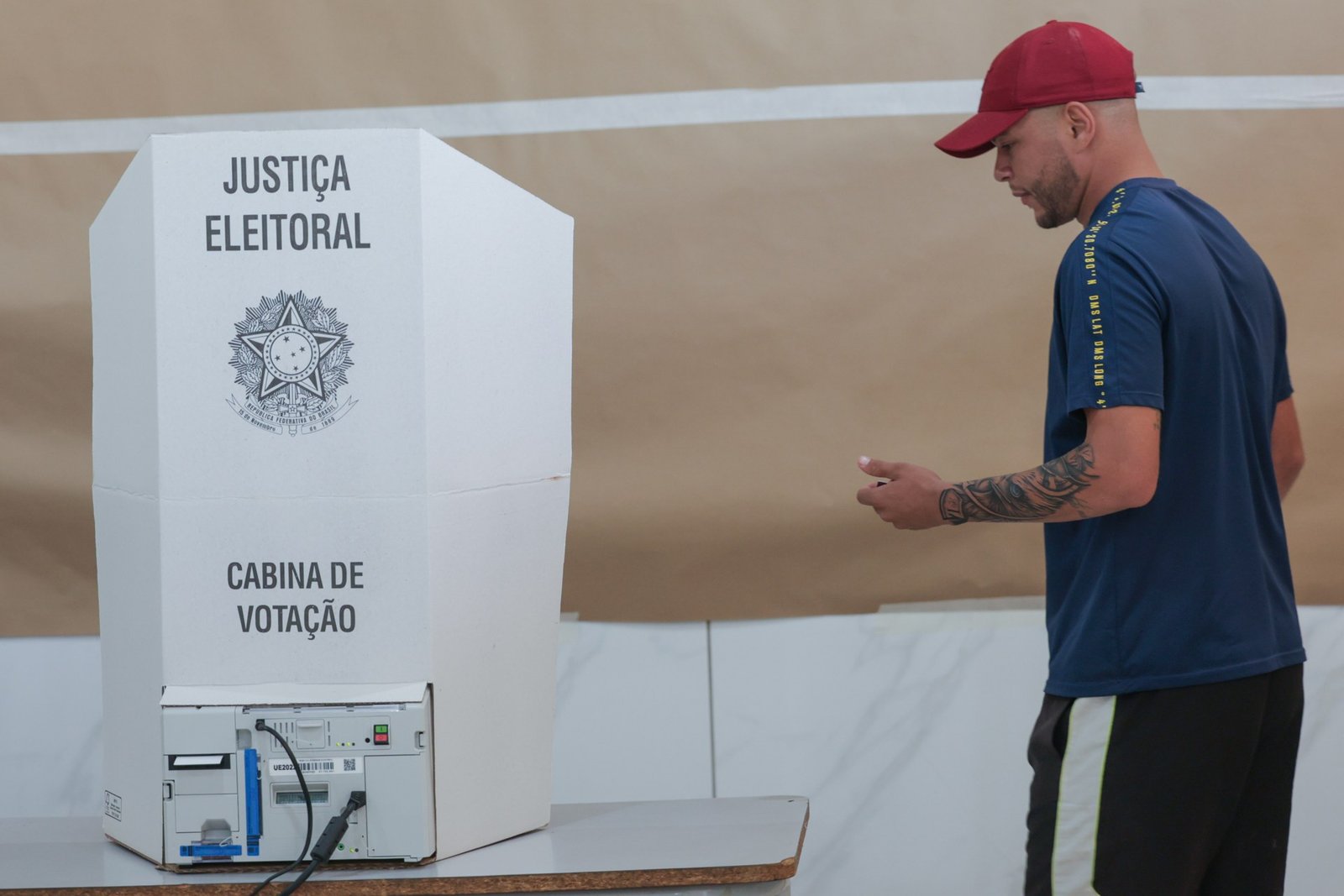 Eleitor de boné vermelho caminha ao lado da cabine de votação da Justiça Eleitoral, com a urna eletrônica sobre a mesa, no primeiro turno