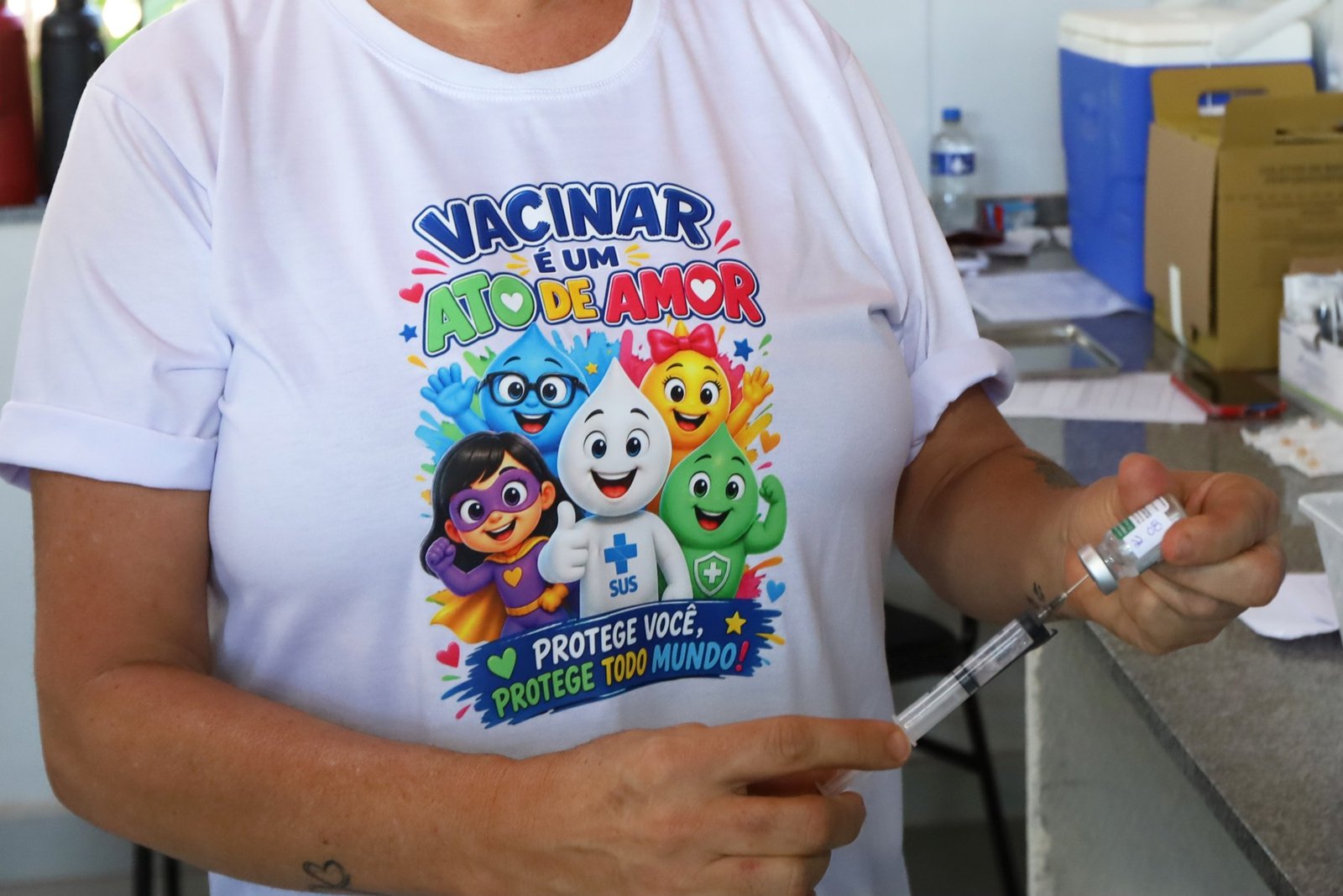Profissional de saúde prepara seringa com vacina em sala de vacinação do DF, com camiseta da campanha do SUS