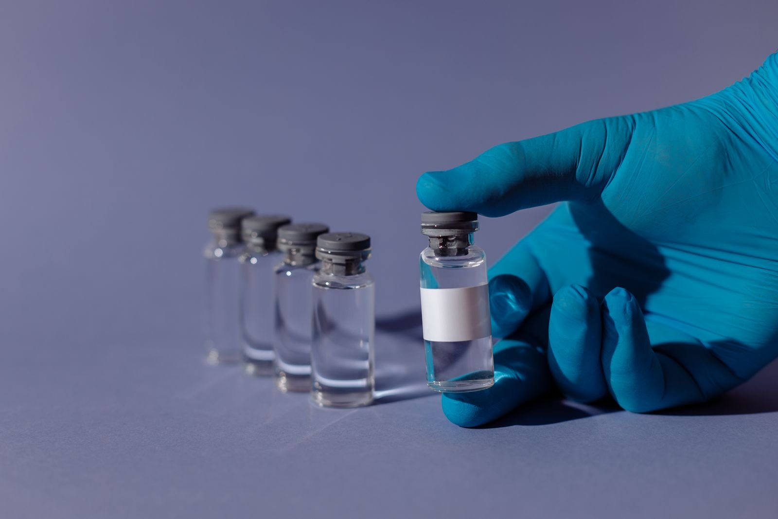 Mão de luva azul segura frasco de vacina ao lado de outros frascos transparentes