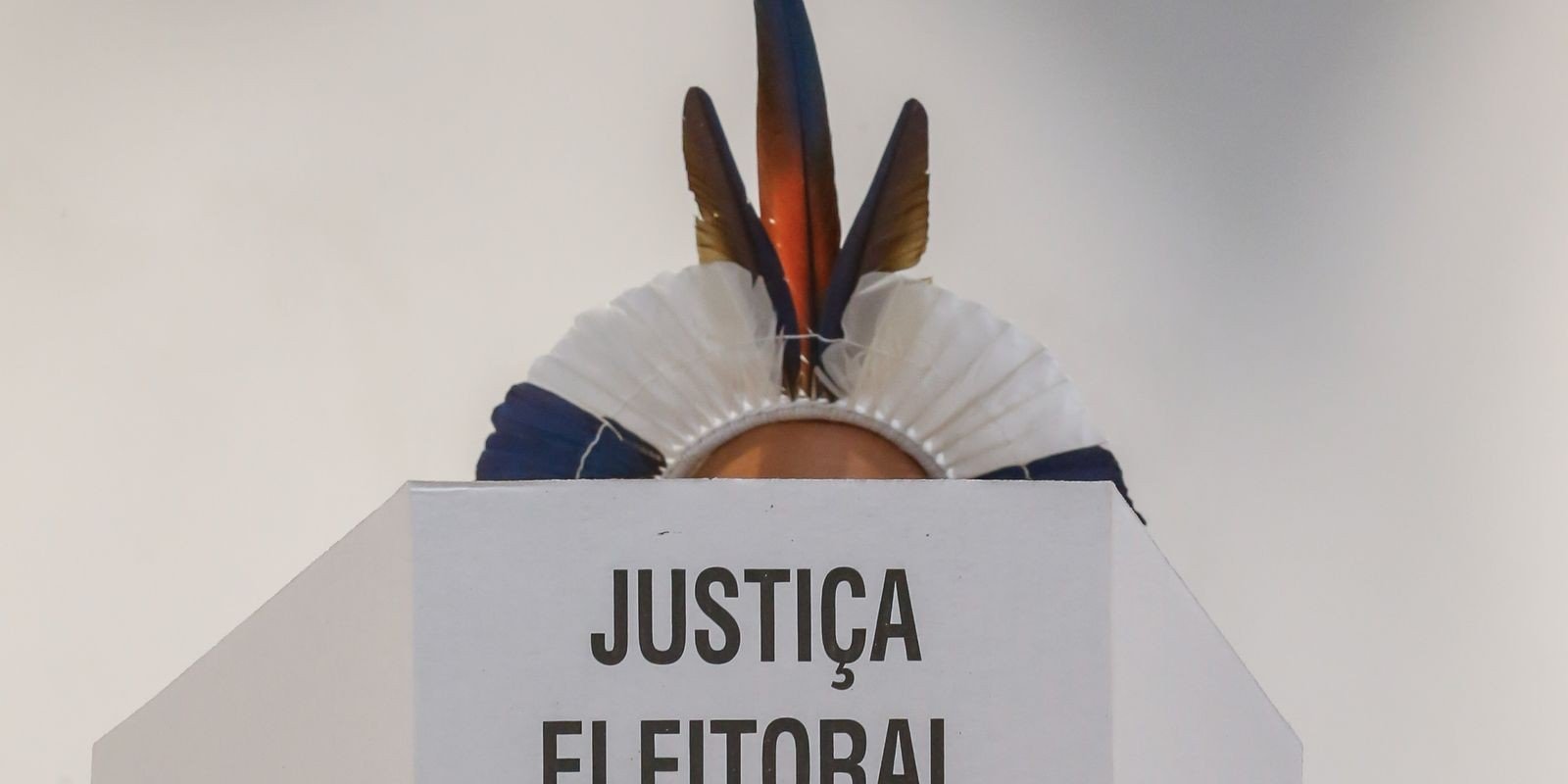 Eleitor indígena com cocar de penas atrás de cabine de votação com a inscrição Justiça Eleitoral