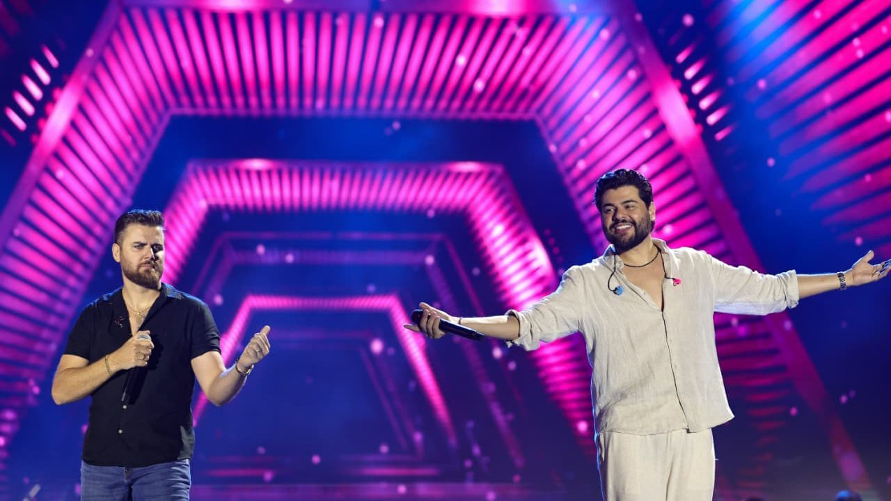 Zé Neto e Cristiano cantam no palco iluminado por luzes rosa e roxas, em show de outra cidade